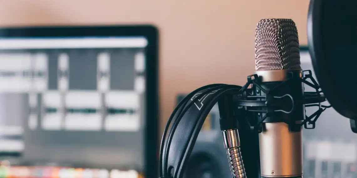 An immersive snapshot of podcasting: A sleek silver condenser microphone takes center stage in a spider stand, with a hint of a noise filter attached. Adjacent, headphones dangle, signaling a recent recording session. The frame expands to reveal a computer screen on the left, showcasing an audio-processing program in action. Audio waves on the screen signify meticulous editing, capturing the essence of the podcast creation process.