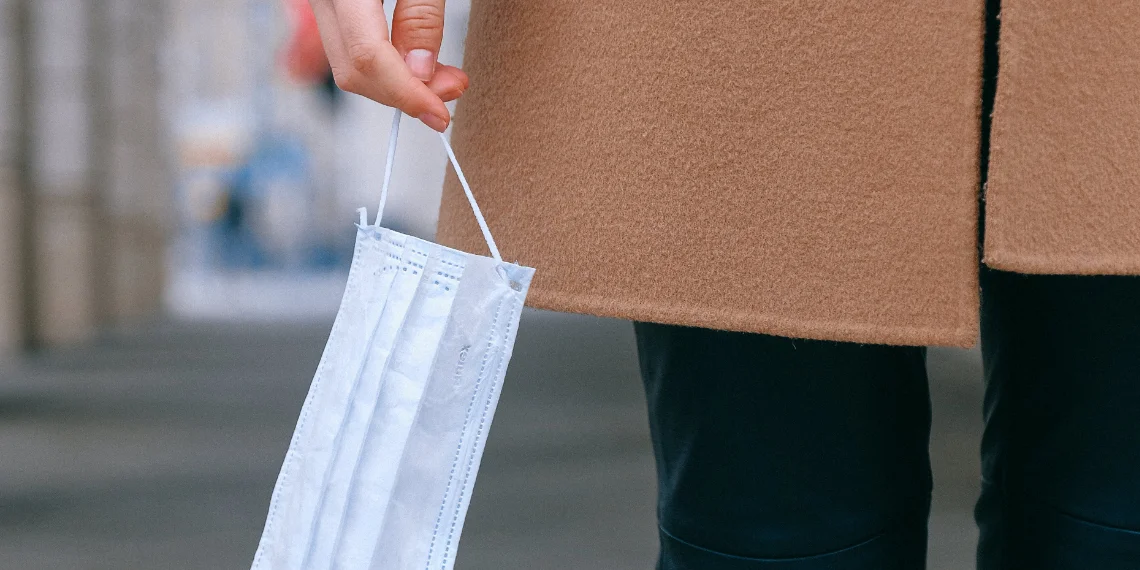 Illustration accompanying a blog post about COVID-19: A woman's lower body is featured, wearing a stylish brown felt coat and black jeans. In her right hand, she holds a typical blue and white surgical face mask by the earstrap, emphasizing the importance of mask-wearing during the pandemic.