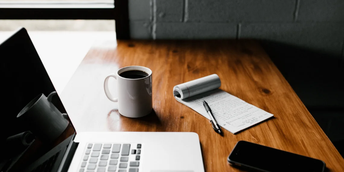 Desk setup with a laptop, coffee cup, notebook, pen, and phone.