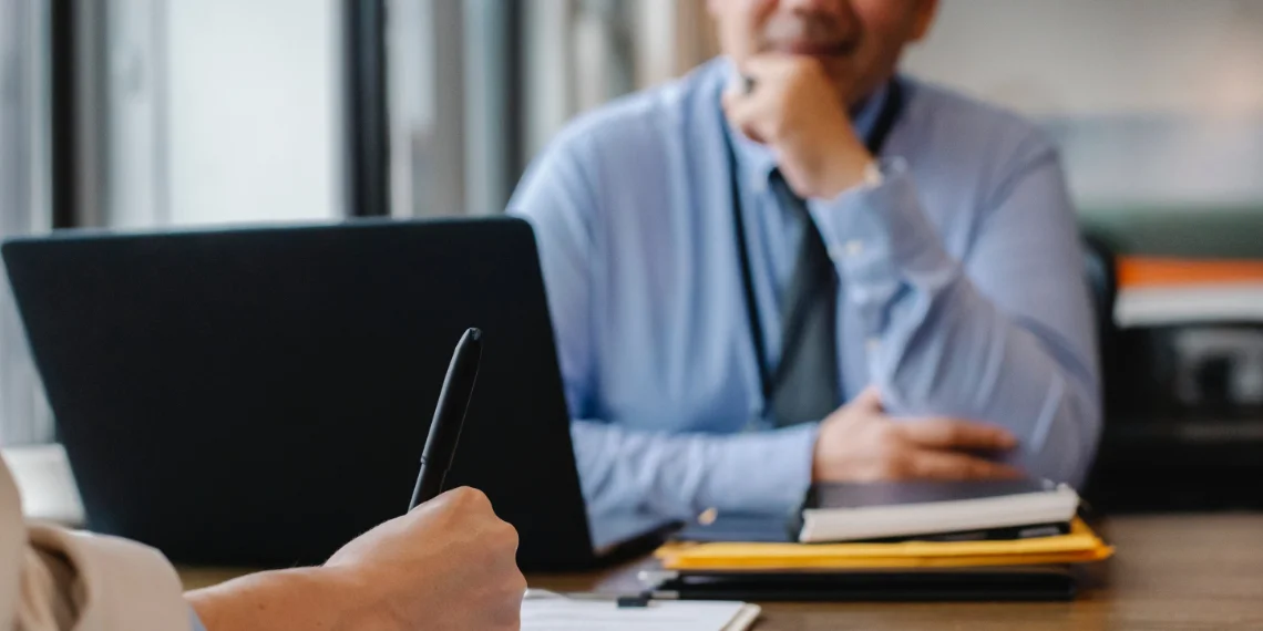 Student interviews for university admissions: A hand holds a pen, writing on a clipboard, while the interviewer attentively evaluates the applicant's profile with folded hands and a thoughtful expression.