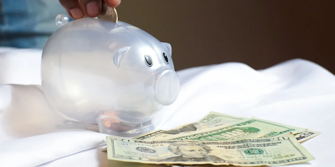 Plastic piggy bank on a desk with three 20-dollar bills in front. Hand, fingertips visible, tossing a quarter into the piggy bank.